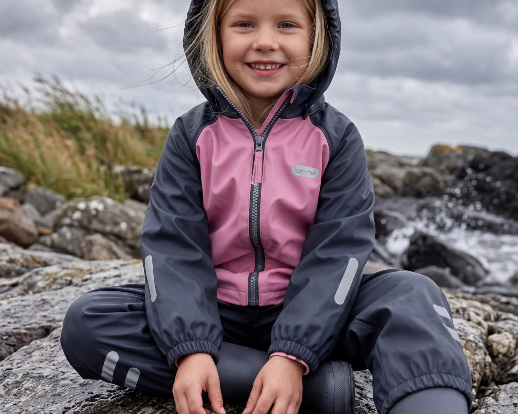 Girl in a pink and navy fleece lined rain set by Lenne, sitting on rocks by the sea