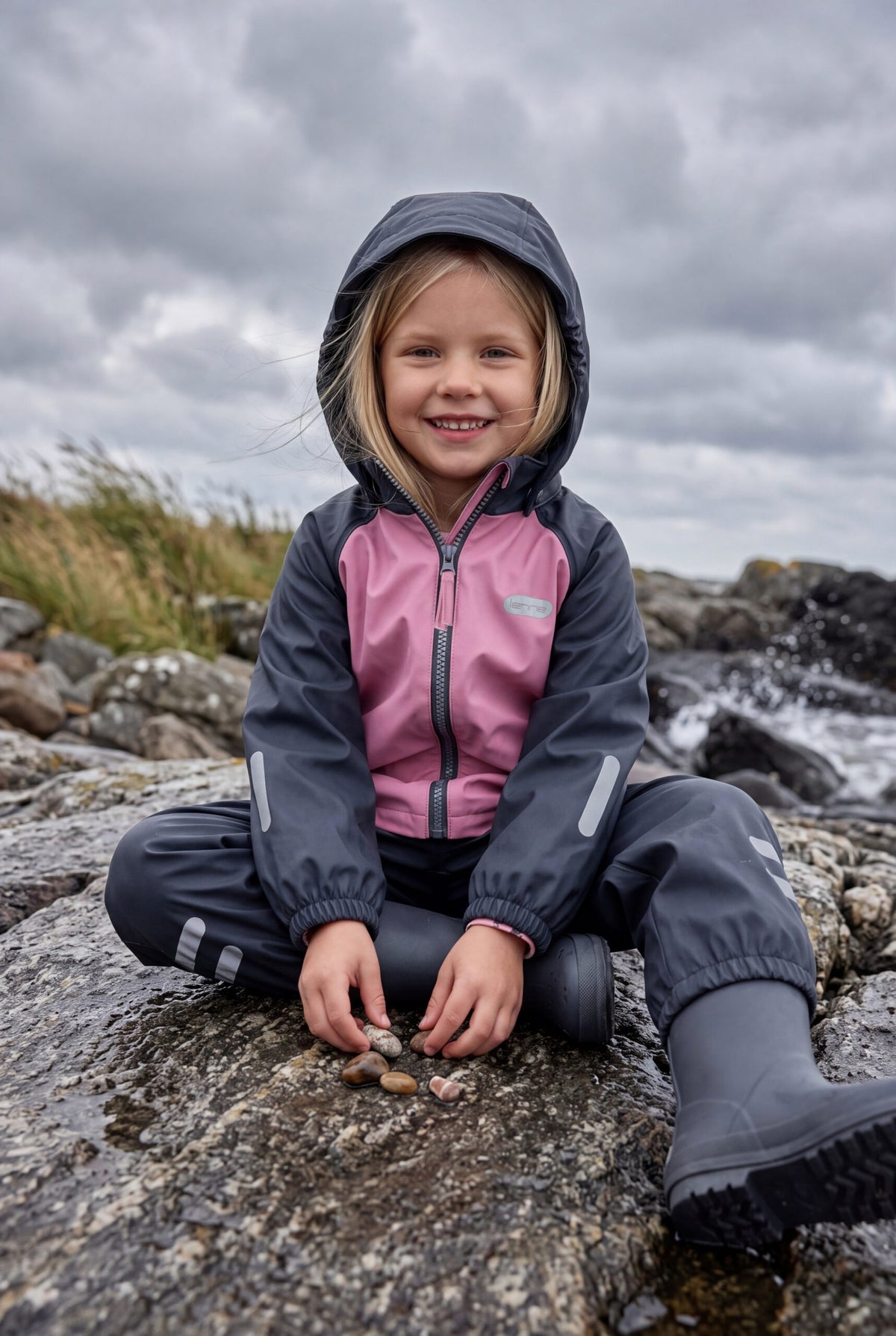 Pige i et lyserødt og marineblåt regnsæt med fleecefor fra Lenne, siddende på klipper ved havet