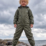 Boy in a green insulated puddle suit by Lenne, standing on rocks by the sea