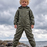 Boy in a green insulated puddle suit by Lenne, standing on rocks by the sea