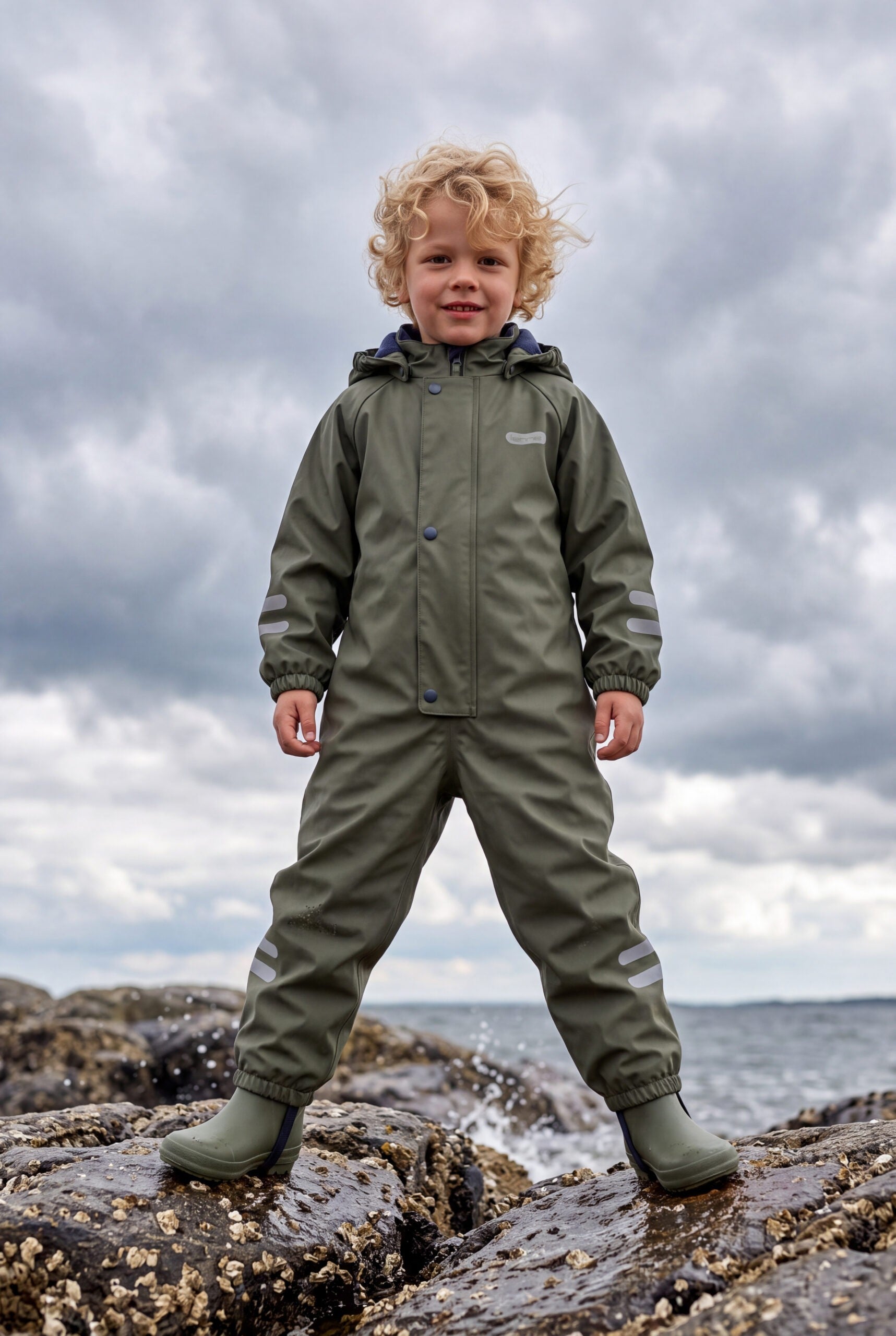 Boy in a green insulated puddle suit by Lenne, standing on rocks by the sea