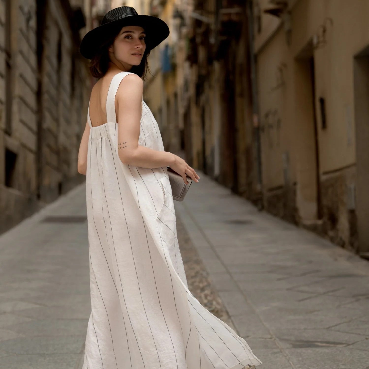 Woman in a black hat wearing the milk white A line linen dress with a grey stripe by Linen Tales, looking back on a stone street