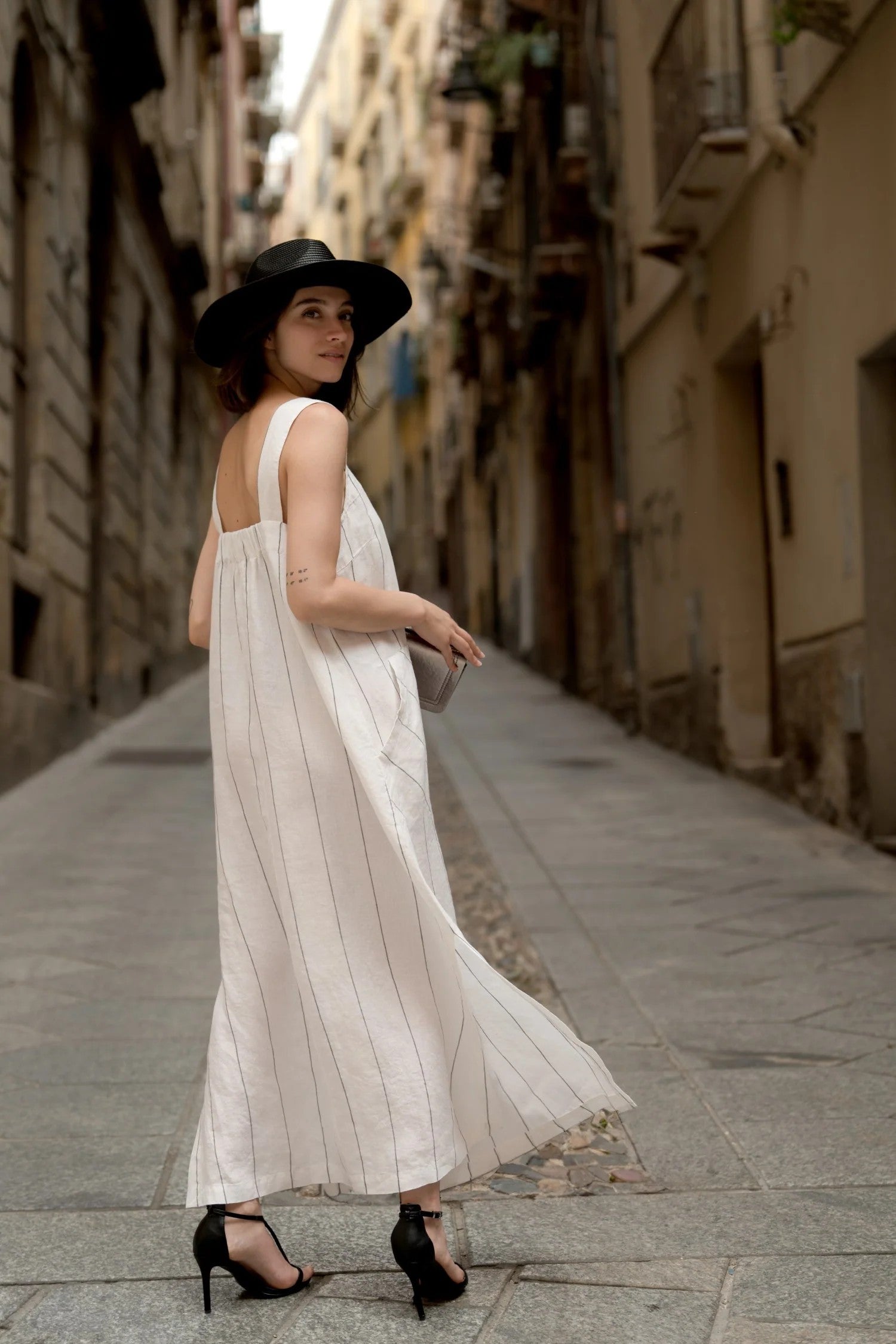 Woman in a black hat wearing the milk white A line linen dress with a grey stripe by Linen Tales, looking back on a stone street