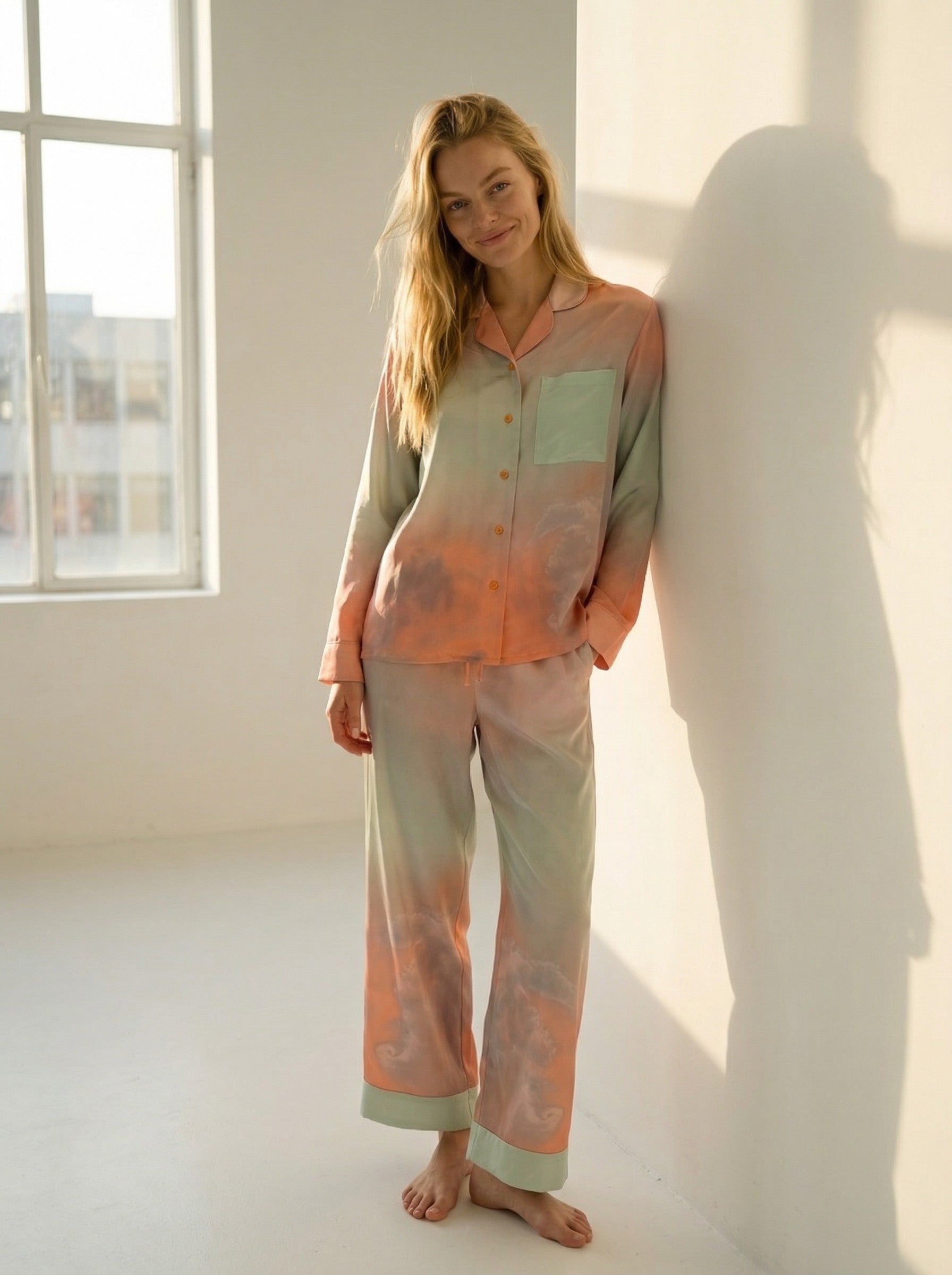 Tæwas Sunset silk pyjamas, button front shirt and drawstring trousers, worn leaning against a wall